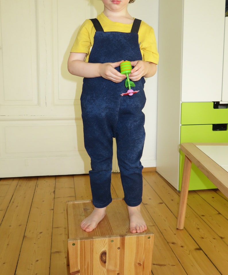 May include: A child wearing a blue denim overall jumpsuit with yellow straps over a yellow short-sleeved shirt. The child is standing on a wooden box.