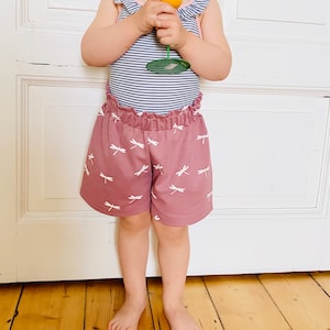 Op de afbeelding: Een kinderoutfit met een gestreepte bodysuit met ruchesmouwen en een stoffige roze short. De short is versierd met een wit libellenpatroon. Het kind houdt een gele speelgoedbloem vast.