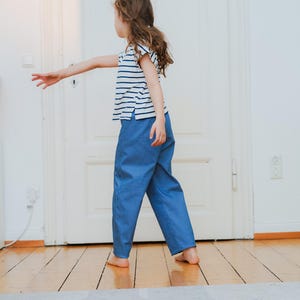 May include: A child wearing a blue and white striped t-shirt and blue pants. The child is walking on a wooden floor. The outfit is casual and comfortable.