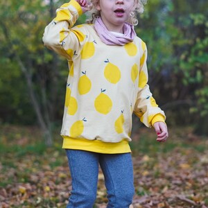 May include: A young person wearing a yellow sweatshirt with a lemon print and blue jeans. The sweatshirt has a yellow band around the bottom.