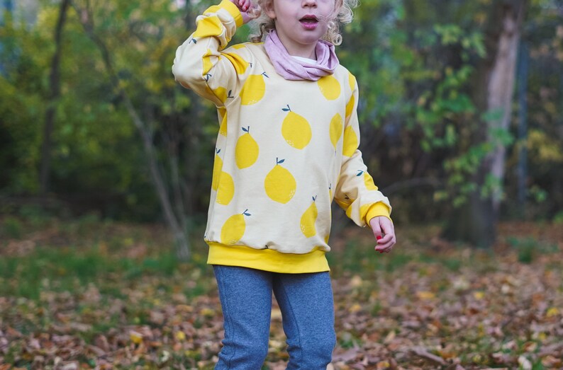May include: A young person wearing a yellow sweatshirt with a lemon print and blue jeans. The sweatshirt has a yellow band around the bottom.