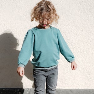 May include: A young person wearing a light blue long-sleeved shirt with a gathered detail at the shoulders and gray jeans.