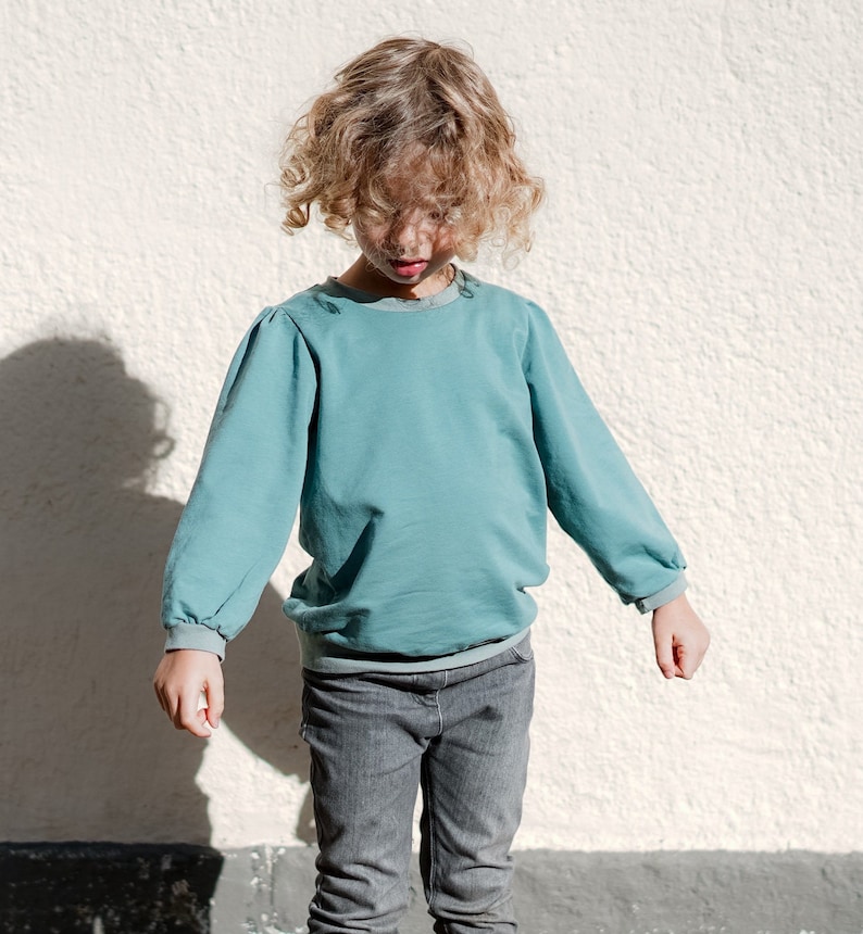 May include: A young person wearing a light blue long-sleeved shirt with a gathered detail at the shoulders and gray jeans.