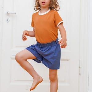 Op de afbeelding: Een kind draagt een oranje t-shirt met witte randen en een blauwe korte broek. Het t-shirt heeft een witte halslijn en manchetten. De korte broek heeft een gerimpelde tailleband. Het kind staat voor een witte deur.