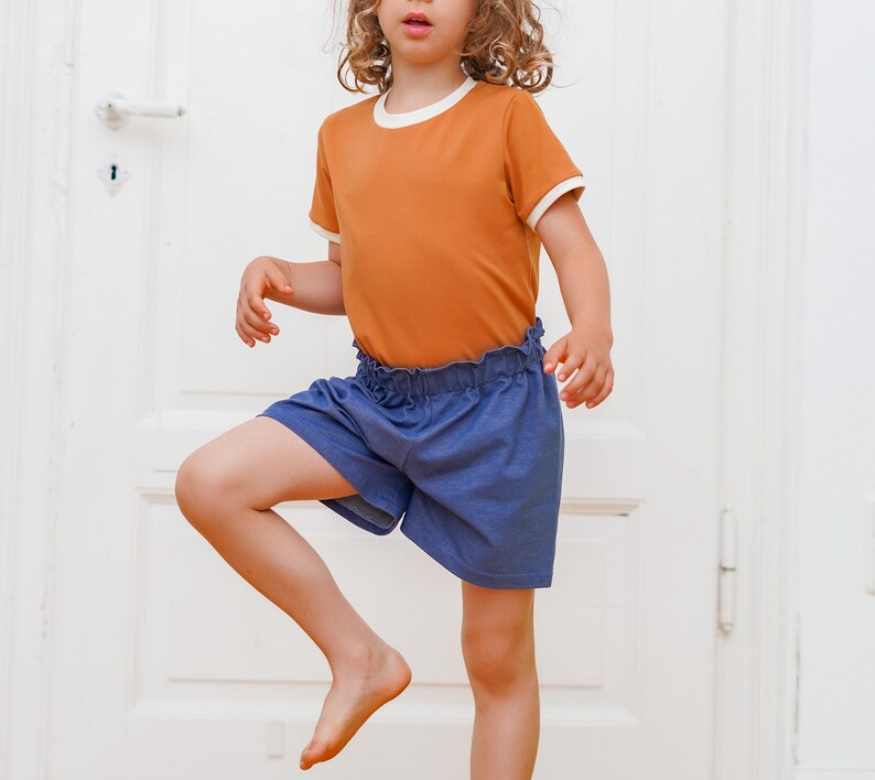 Op de afbeelding: Een kind draagt een oranje t-shirt met witte randen en een blauwe korte broek. Het t-shirt heeft een witte halslijn en manchetten. De korte broek heeft een gerimpelde tailleband. Het kind staat voor een witte deur.