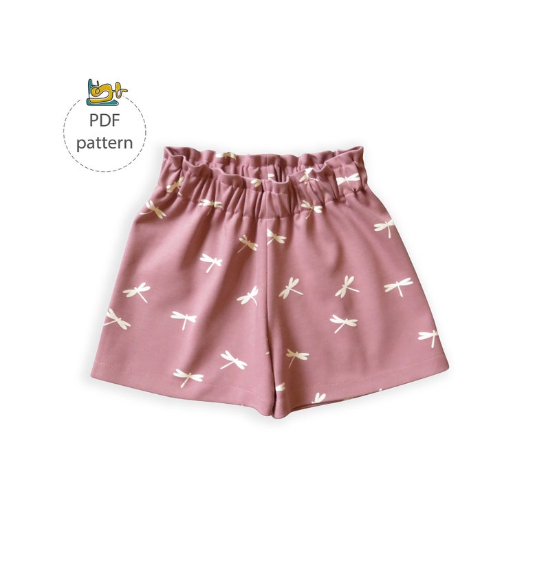 Op de afbeelding: Zachtroze shorts met een wit libellenpatroon en een elastische tailleband. De shorts zijn gemaakt van een zachte stof. De afbeelding bevat ook een kleine afbeelding met de tekst "PDF pattern".
