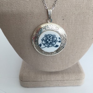 May include: Silver-tone locket necklace with a floral design. The round pendant features a white ceramic insert with a blue floral pattern. The necklace is displayed on a beige linen-textured surface.