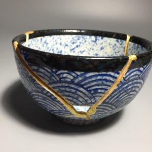 May include: A ceramic bowl featuring a blue and white wave pattern on the exterior and a speckled blue and white interior. The bowl has a dark rim and is repaired with gold-colored lacquer lines that highlight the cracks across the surface.