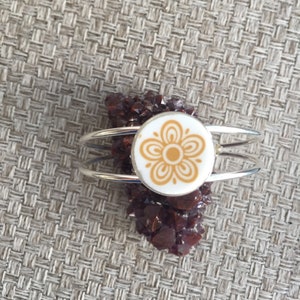 May include: A silver bangle bracelet with a white ceramic charm featuring a gold floral design. The charm is set on a bed of red crystals.