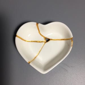 May include: A white heart-shaped ceramic dish featuring a repair technique with visible gold-colored lines running across the surface. The dish has a smooth, glossy finish and a small triangular gap where the gold lines intersect in the center.