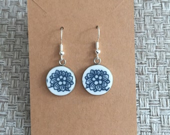 May include: A pair of round earrings with a white background and a blue floral design. The earrings are attached to silver-colored hooks and are displayed on a brown card.