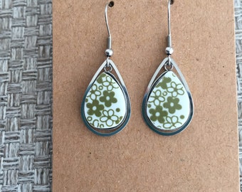 May include: A pair of teardrop earrings with a floral design. The earrings feature a silver-colored frame and a white background with olive green flowers. The earrings are displayed on a brown card.