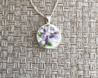 May include: A silver chain necklace with a round pendant featuring a floral design of purple and yellow flowers on a white background.