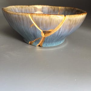 May include: A ceramic bowl featuring a kintsugi repair with gold-colored lacquer filling the cracks. The bowl has a gradient glaze transitioning from a light tan at the rim to a soft blue at the base, with a smooth, rounded shape.