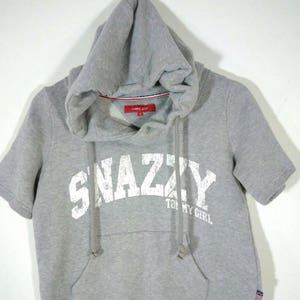 May include: A gray short-sleeved hoodie with a drawstring hood and a front pocket. The word "SNAZZY" is printed across the chest in white, with "TOMMY GIRL" below. The hoodie is hanging against a white background.