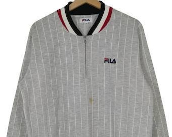 May include: A gray and white striped long-sleeve shirt with a black and red collar. The shirt has a zipper closure and the word "FILA" is embroidered on the front.