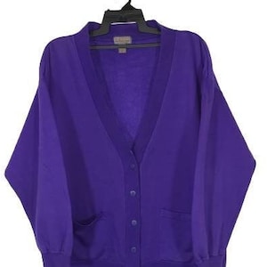Può includere: Un cardigan viola con scollo a V, maniche lunghe e chiusura con bottoni. Ha due tasche sul davanti e un orlo a coste.