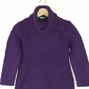 May include: A purple turtleneck sweater with a chunky knit texture. The sweater is hanging on a wooden hanger.