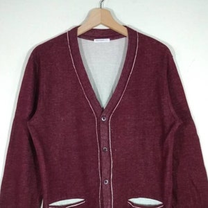 May include: A maroon cardigan with white trim around the collar, pockets, and button placket. The cardigan has a V-neck and long sleeves.