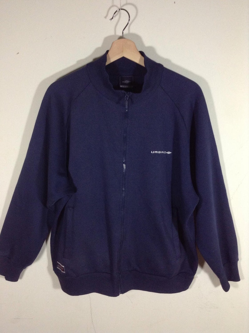 Pu&ograve; includere: Una felpa blu navy con zip e logo "UMBRO" bianco sul davanti.