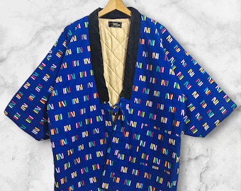 Puede incluir: Chaqueta azul con un colorido estampado geométrico. La chaqueta tiene un cuello negro y un forro acolchado beige. Cuelga de una percha de madera.