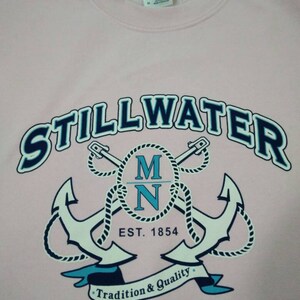 Puede incluir: Sudadera rosa con un gr&aacute;fico azul marino y blanco de dos anclas cruzadas con cuerda, el texto "STILLWATER MN EST. 1854" y una pancarta que dice "Tradici&oacute;n y Calidad".