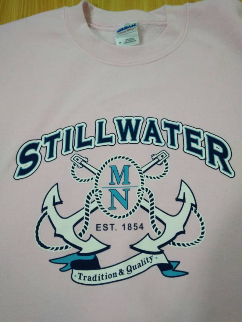 Puede incluir: Sudadera rosa con un gr&aacute;fico azul marino y blanco de dos anclas cruzadas con cuerda, el texto "STILLWATER MN EST. 1854" y una pancarta que dice "Tradici&oacute;n y Calidad".