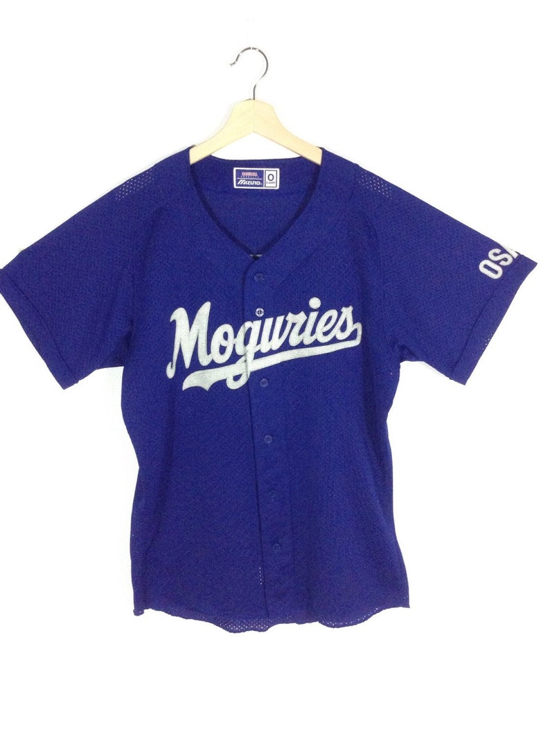 May include: A royal blue baseball jersey with white mesh fabric. The jersey has a white "Moguries" logo on the front and a white "OSA" logo on the right sleeve. The jersey has a button-up front.