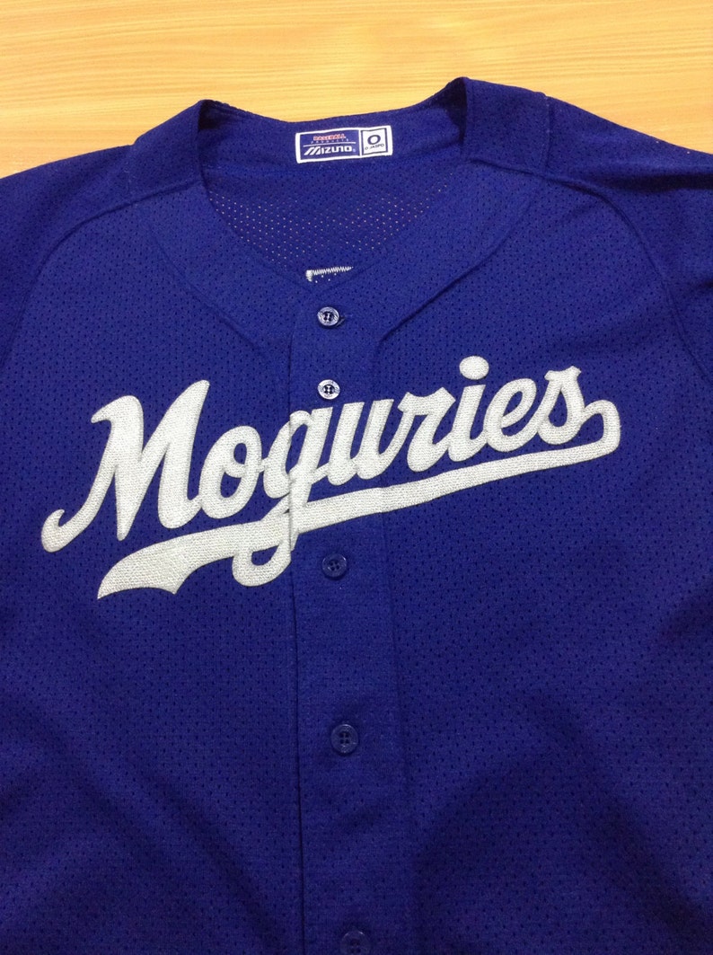 May include: A royal blue baseball jersey with white lettering that reads "Moguries". The jersey has a button-up front and a mesh fabric.