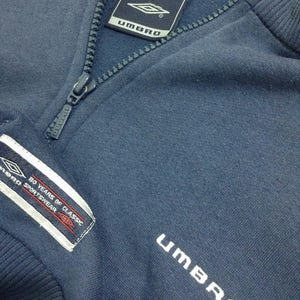 Pu&ograve; includere: Una felpa blu navy con cerniera che presenta la scritta "UMBRO" ricamata sul davanti e un'etichetta bianca con il testo "80 YEARS OF CLASSIC SPORTSWEAR" sul lato sinistro.