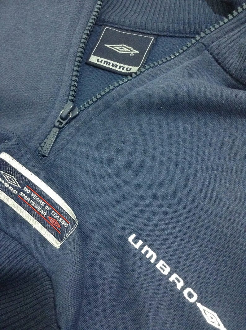 Pu&ograve; includere: Una felpa blu navy con cerniera che presenta la scritta "UMBRO" ricamata sul davanti e un'etichetta bianca con il testo "80 YEARS OF CLASSIC SPORTSWEAR" sul lato sinistro.
