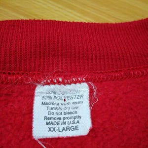May include: A white tag with black text attached to a red sweatshirt. The tag says "50% Polyester, Machine wash warm, Tumble dry low, Do not bleach, Remove promptly, Made in U.S.A., XX-Large."