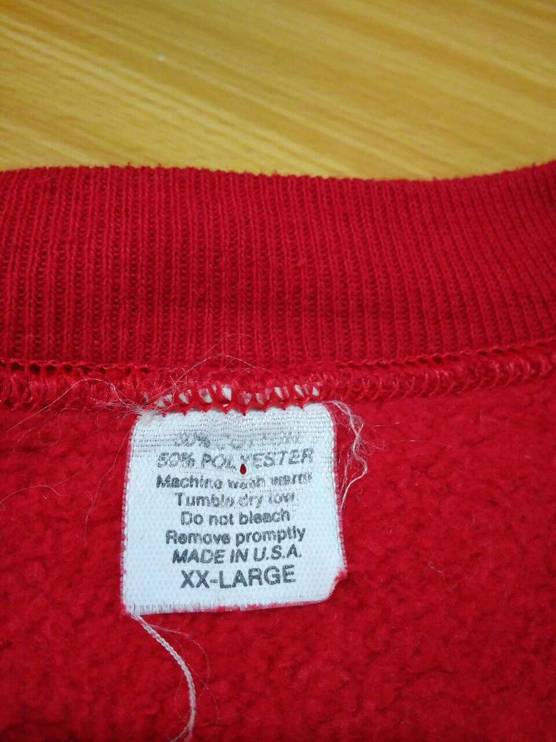 May include: A white tag with black text attached to a red sweatshirt. The tag says "50% Polyester, Machine wash warm, Tumble dry low, Do not bleach, Remove promptly, Made in U.S.A., XX-Large."