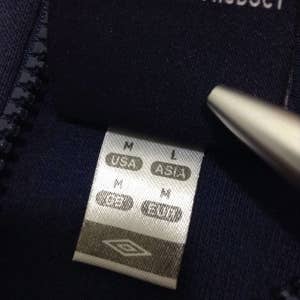 Pu&ograve; includere: Primo piano di una giacca blu navy con cerniera che presenta un'etichetta bianca con la scritta "OFFICIAL UMBRO PRODUCT" e una tabella delle taglie con le taglie "USA" e "ASIA".