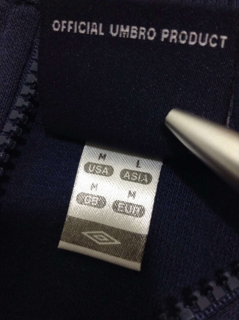 Pu&ograve; includere: Primo piano di una giacca blu navy con cerniera che presenta un'etichetta bianca con la scritta "OFFICIAL UMBRO PRODUCT" e una tabella delle taglie con le taglie "USA" e "ASIA".