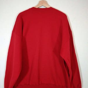 May include: A red sweatshirt with a crew neck and long sleeves. The sweatshirt is hanging on a wooden hanger.