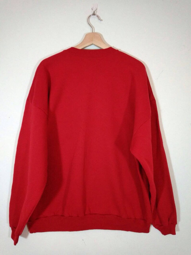 May include: A red sweatshirt with a crew neck and long sleeves. The sweatshirt is hanging on a wooden hanger.