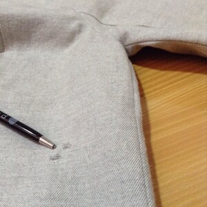 May include: A close-up of a grey hooded sweatshirt with a small hole in the fabric. A black pen is lying on the fabric.