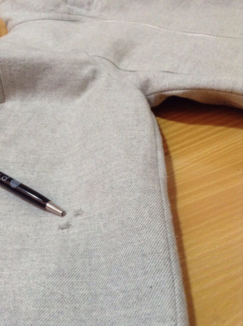 May include: A close-up of a grey hooded sweatshirt with a small hole in the fabric. A black pen is lying on the fabric.