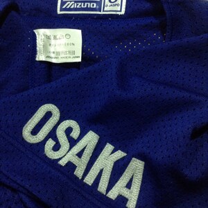 May include: Blue baseball jersey with a white "OSAKA" logo on the sleeve. The jersey has a mesh fabric and a Mizuno label with "O JASPO" on the collar.