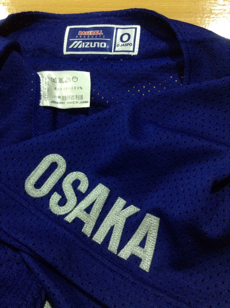 May include: Blue baseball jersey with a white "OSAKA" logo on the sleeve. The jersey has a mesh fabric and a Mizuno label with "O JASPO" on the collar.