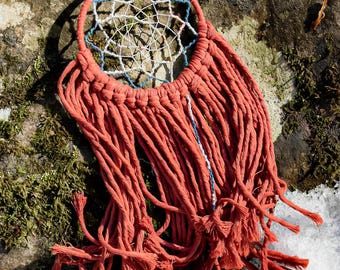 May include: A red and blue dreamcatcher with a white web and red fringe hanging on a rock covered in moss.