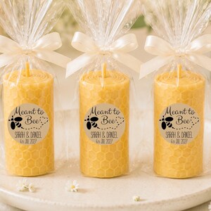 May include: Three cylindrical yellow beeswax candles with a honeycomb texture, each wrapped in clear plastic and tied with a white satin ribbon. A circular sticker on each candle features a black bee graphic and the text Meant to Bee, Sarah & Daniel, 4th Jul 2027.