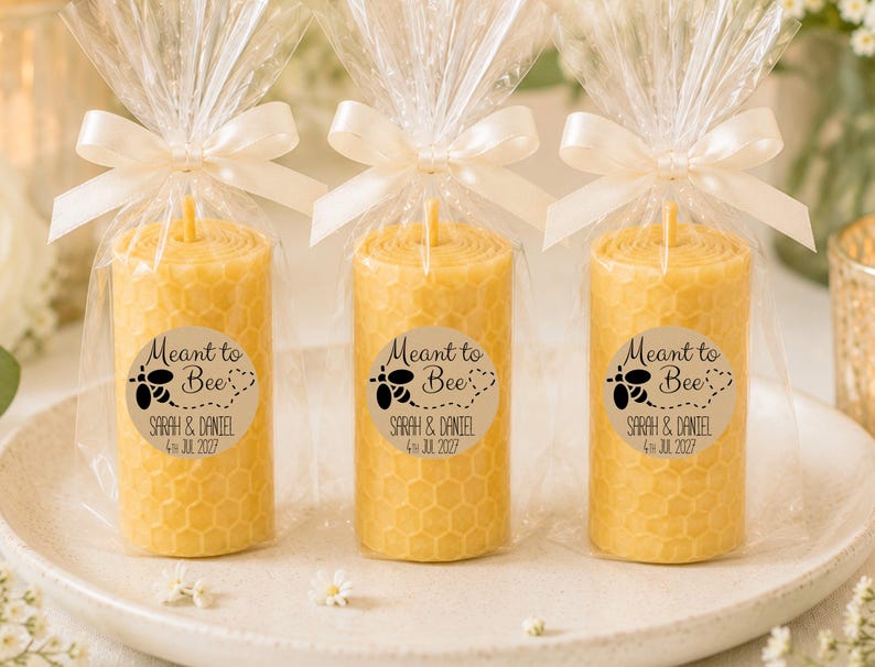May include: Three cylindrical yellow beeswax candles with a honeycomb texture, each wrapped in clear plastic and tied with a white satin ribbon. A circular sticker on each candle features a black bee graphic and the text Meant to Bee, Sarah & Daniel, 4th Jul 2027.