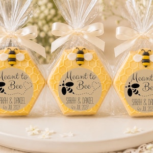 May include: Three hexagonal yellow soaps shaped like honeycombs, each topped with a small bee figure. They are wrapped in clear plastic with a white ribbon and a circular label reading Meant to Bee, Sarah & Daniel, 4th Jul 2027.