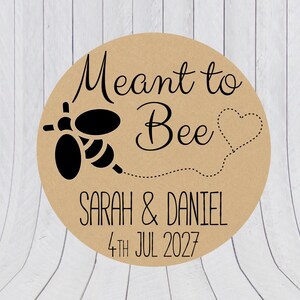 May include: A round tan sticker featuring a black graphic of a bee with a dotted line trail forming a heart shape. The text reads Meant to Bee, Sarah & Daniel, and 4th Jul 2027 in black lettering. The design is centered on a circular kraft paper background.