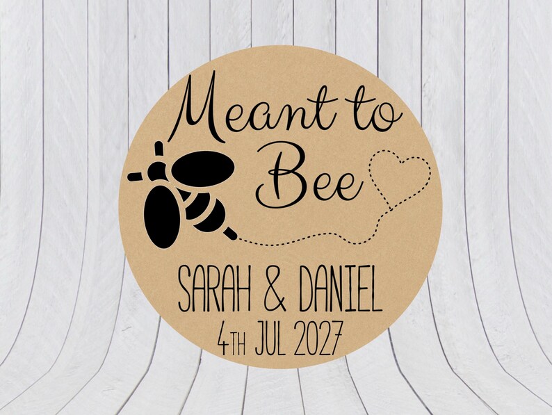 May include: A round tan sticker featuring a black graphic of a bee with a dotted line trail forming a heart shape. The text reads Meant to Bee, Sarah & Daniel, and 4th Jul 2027 in black lettering. The design is centered on a circular kraft paper background.