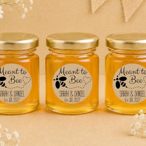 May include: Three clear glass jars filled with golden honey, each topped with a gold metal lid. A circular beige label on each jar features a black bee graphic, a dotted heart trail, and the text Meant to Bee, Sarah & Daniel, 4th Jul 2027.