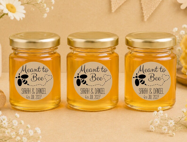 May include: Three clear glass jars filled with golden honey, each topped with a gold metal lid. A circular beige label on each jar features a black bee graphic, a dotted heart trail, and the text Meant to Bee, Sarah & Daniel, 4th Jul 2027.