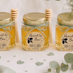 May include: Three hexagonal glass jars filled with golden honey, each topped with a gold lid and a wooden honey dipper. A light green twine bow secures the dipper. A round label reads "Meant to Bee" with a bee graphic and "SARAH & DANIEL 4TH JUL 2027".